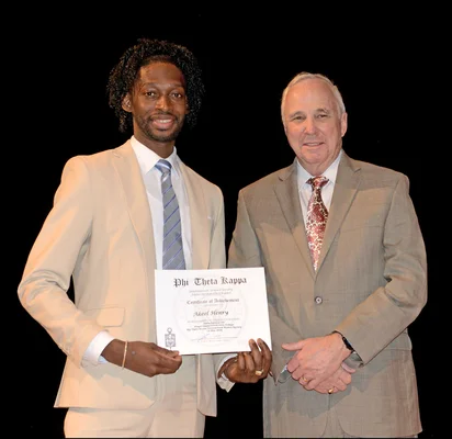 Two men in suits look forward smiling as one holds an honor society certificate