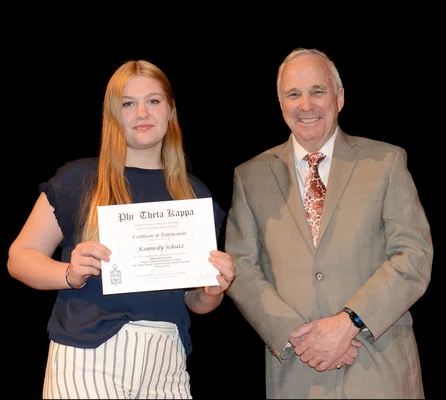 A young woman holds and honor society certificate as she poses with a man in a suit