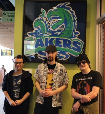 Three men facing the camera posing in front of the LEGO mosaic at Finger Lakes Community College