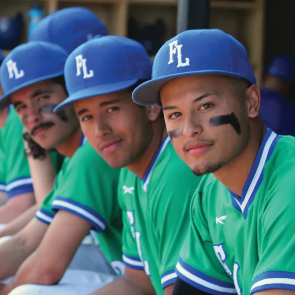 The men's baseball team sit in a dugout, ready for the game to start.